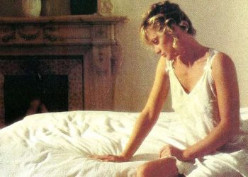 Woman in a nightgown sitting thoughtfully on a bed in a softly lit bedroom, suggesting discomfort or concern.