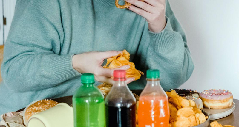 Person eating fast food and sugary snacks, surrounded by fizzy drinks and pastries, representing binge eating disorder (BED) behaviour.