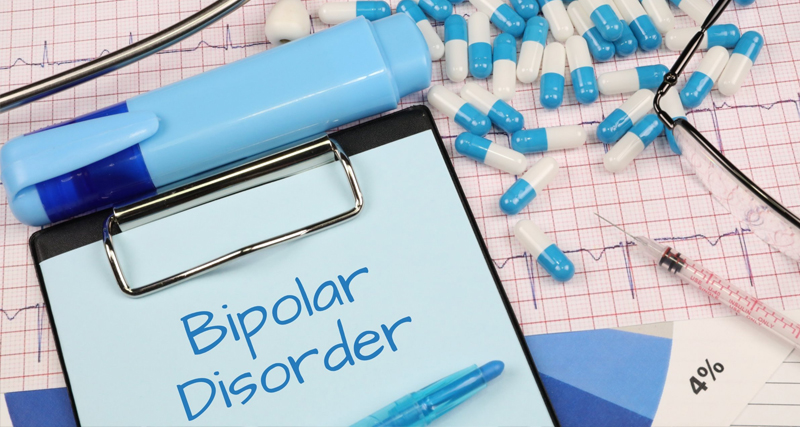 Medical chart with the words "Bipolar Disorder," surrounded by blue and white capsules, syringe, and ECG printout.