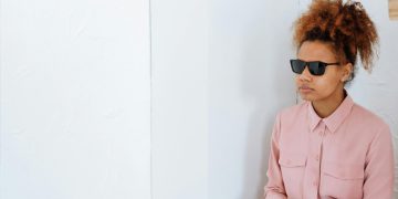 Woman wearing sunglasses indoors, possibly blind or visually impaired, seated against a white background.
