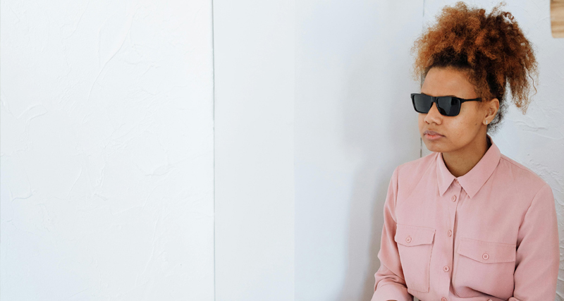 Woman wearing sunglasses indoors, possibly blind or visually impaired, seated against a white background.