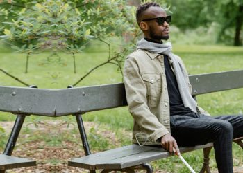 Blind man sitting on a park bench wearing sunglasses and holding a white cane.
