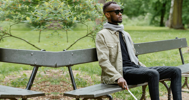 Blind man sitting on a park bench wearing sunglasses and holding a white cane.