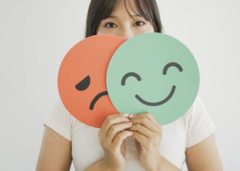 Woman holding smiling and frowning face icons in front of her mouth