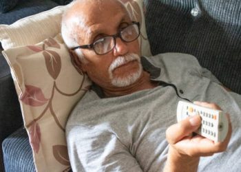 Elderly man lying on a couch with a remote control, symbolising inactivity as a risk factor for coronary heart disease.
