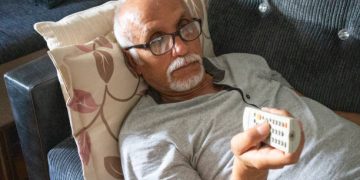 Elderly man lying on a couch with a remote control, symbolising inactivity as a risk factor for coronary heart disease.