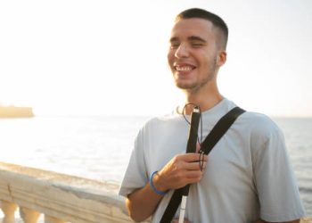Young man with visual impairment smiling while standing near the ocean, holding a white cane