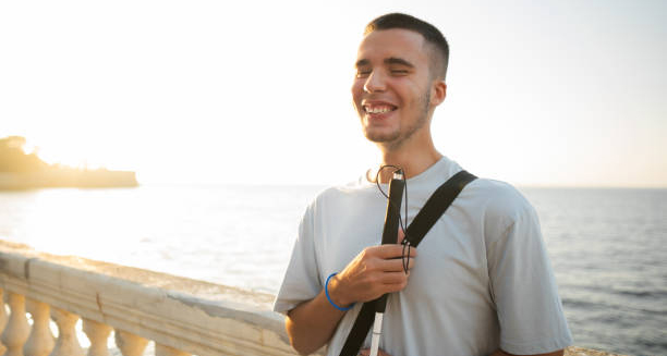 Young man with visual impairment smiling while standing near the ocean, holding a white cane