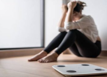 Woman sitting distressed near a weighing scale, symbolising psychological causes of eating disorders