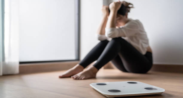 Woman sitting distressed near a weighing scale, symbolising psychological causes of eating disorders