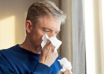 Middle-aged man blowing his nose while standing by a window