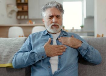 Elderly man clutching his chest in discomfort while sitting on a couch, indicating heart-related complications.