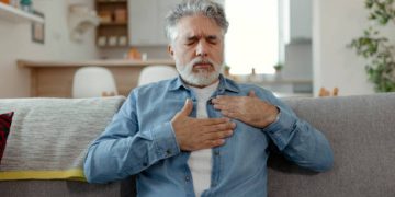 Elderly man clutching his chest in discomfort while sitting on a couch, indicating heart-related complications.