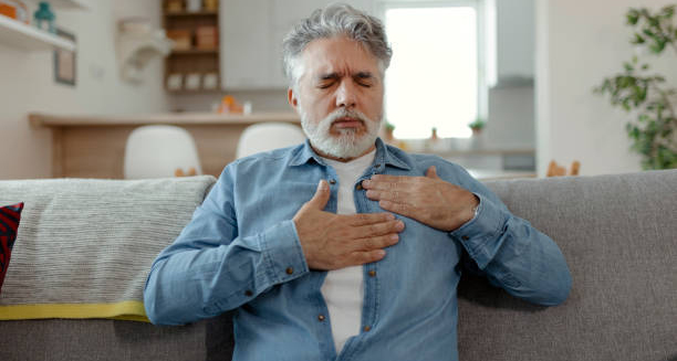 Elderly man clutching his chest in discomfort while sitting on a couch, indicating heart-related complications.
