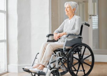 Elderly woman in a wheelchair looking out a window, symbolising mobility challenges related to corticobasal degeneration.