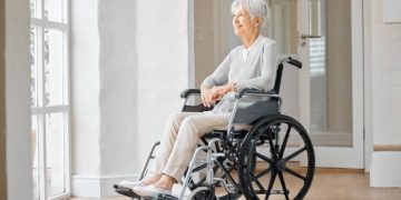 Elderly woman in a wheelchair looking out a window, symbolising mobility challenges related to corticobasal degeneration.