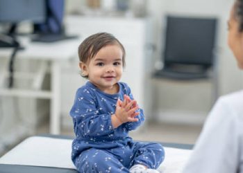 Smiling toddler in pyjamas sitting on a medical exam table, interacting with a doctor, symbolising recovery from craniosynostosis.