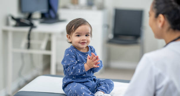 Smiling toddler in pyjamas sitting on a medical exam table, interacting with a doctor, symbolising recovery from craniosynostosis.