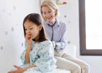 Young girl coughing while being examined by a smiling healthcare provider, symbolising complications and recovery from croup.