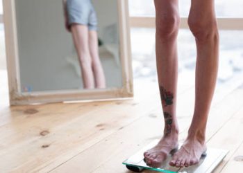 Thin person standing on scale with distorted reflection in mirror