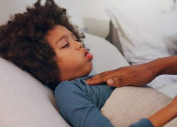 Young child lying in bed with laboured breathing while a caregiver places a comforting hand on their chest, suggesting symptoms of croup.