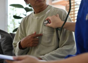 Doctor using a stethoscope to examine an elderly man’s chest, checking for signs of coronary heart disease.