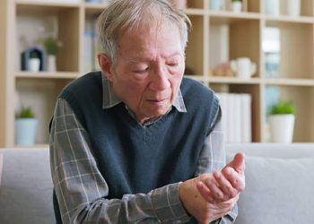 Elderly man sitting on a couch, holding and examining his hand, indicating motor symptoms of corticobasal degeneration.