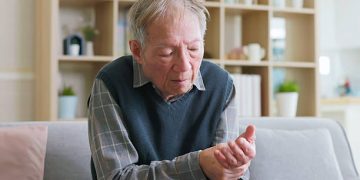 Elderly man sitting on a couch, holding and examining his hand, indicating motor symptoms of corticobasal degeneration.