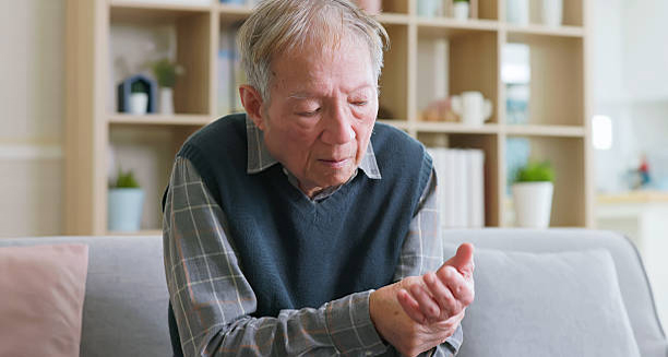 Elderly man sitting on a couch, holding and examining his hand, indicating motor symptoms of corticobasal degeneration.
