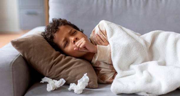 Young child lying on a couch under a blanket, coughing with tissues nearby, showing signs of respiratory illness such as croup.