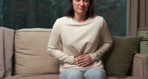 Woman clutching her abdomen in pain while sitting on a couch, showing signs of digestive distress.
