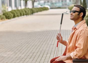 Blind man with a cane sitting on a bench wearing sunglasses in a city setting
