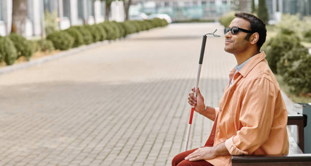 Blind man with a cane sitting on a bench wearing sunglasses in a city setting