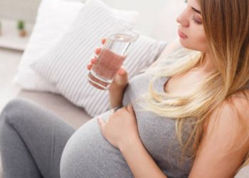 Pregnant woman holding her belly and drinking a glass of water, possibly experiencing symptoms of diabetes in pregnancy
