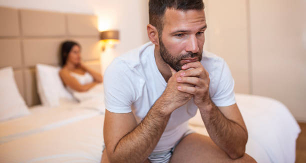 Distressed man sitting on bed with partner in the background, showing symptoms of erectile dysfunction