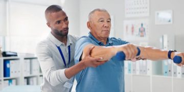 Healthcare professional assisting an elderly man with physical therapy exercises, supporting treatment for corticobasal degeneration.