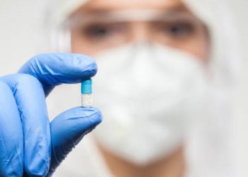 Medical professional in protective gear holding a blue and white capsule between gloved fingers.