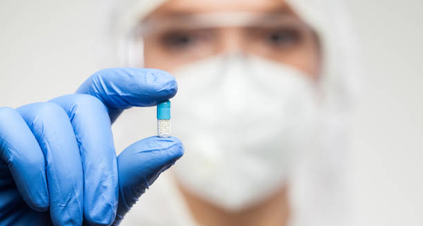 Medical professional in protective gear holding a blue and white capsule between gloved fingers.