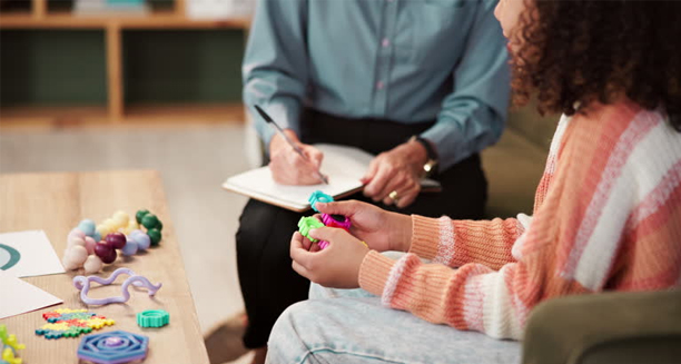 Child engaging in therapy with a psychologist taking notes in the background.