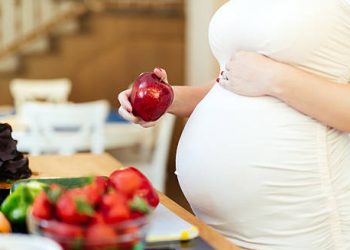 Pregnant woman holding apple with fresh vegetables on table