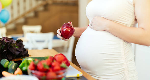 Pregnant woman holding apple with fresh vegetables on table