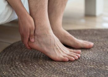 Person holding their ankle in discomfort while sitting on the edge of a bed