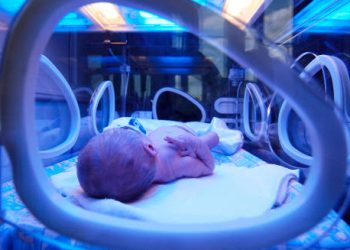 Newborn baby lying in an incubator, viewed through a window, symbolising complications of flat head syndrome