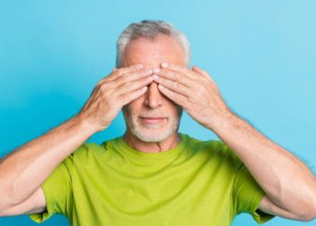 Elderly man covering his eyes with hands on blue background