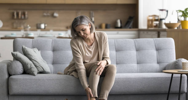 Elderly woman holding leg in pain while seated on a couch