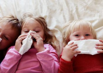 Three young children lying on a bed with tissues, showing cold or flu symptoms
