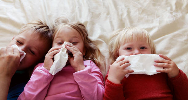 Three young children lying on a bed with tissues, showing cold or flu symptoms