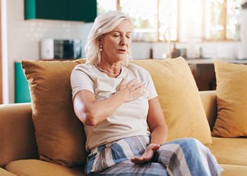 Older woman holding chest in discomfort while sitting on sofa, showing possible heart block complications