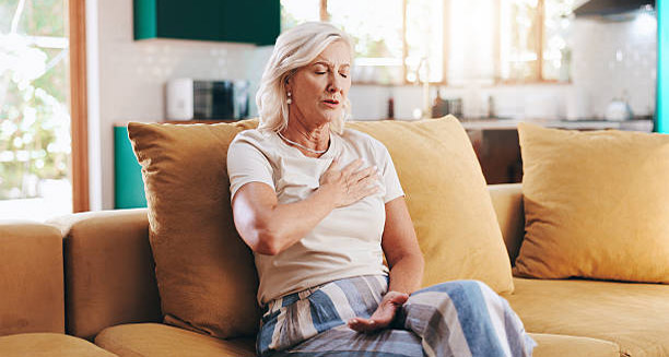 Older woman holding chest in discomfort while sitting on sofa, showing possible heart block complications
