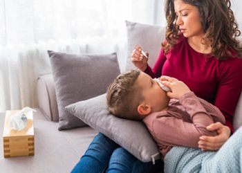 Mother caring for sick child lying on sofa with tissues and thermometer.